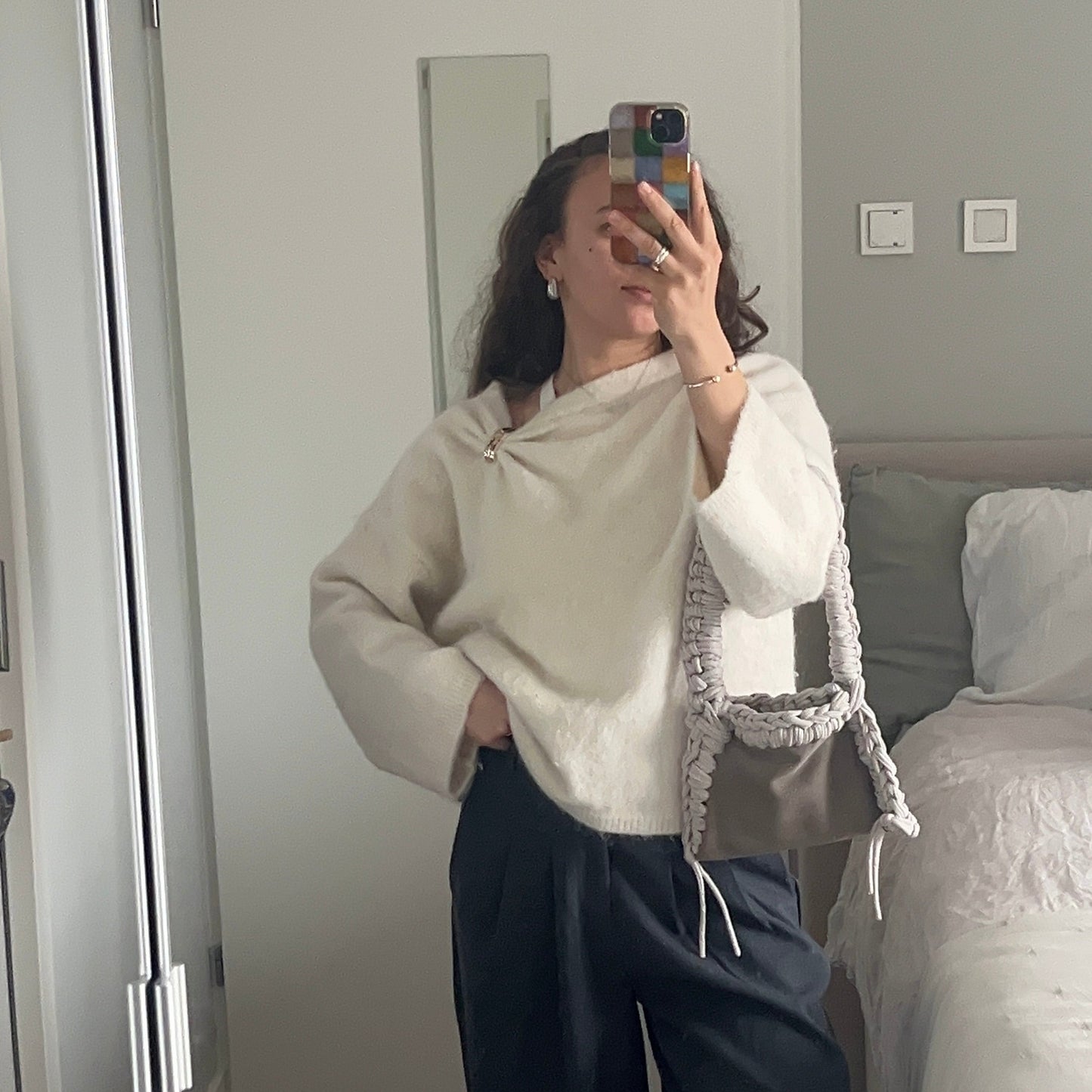 Person taking a mirror selfie wearing a beige sweater and dark pants in a bedroom.