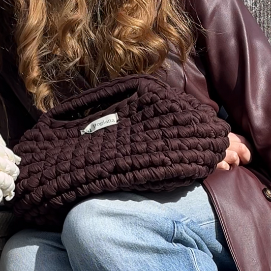 Person wearing a brown leather jacket, dark sweater, and blue jeans sitting on a concrete ledge.