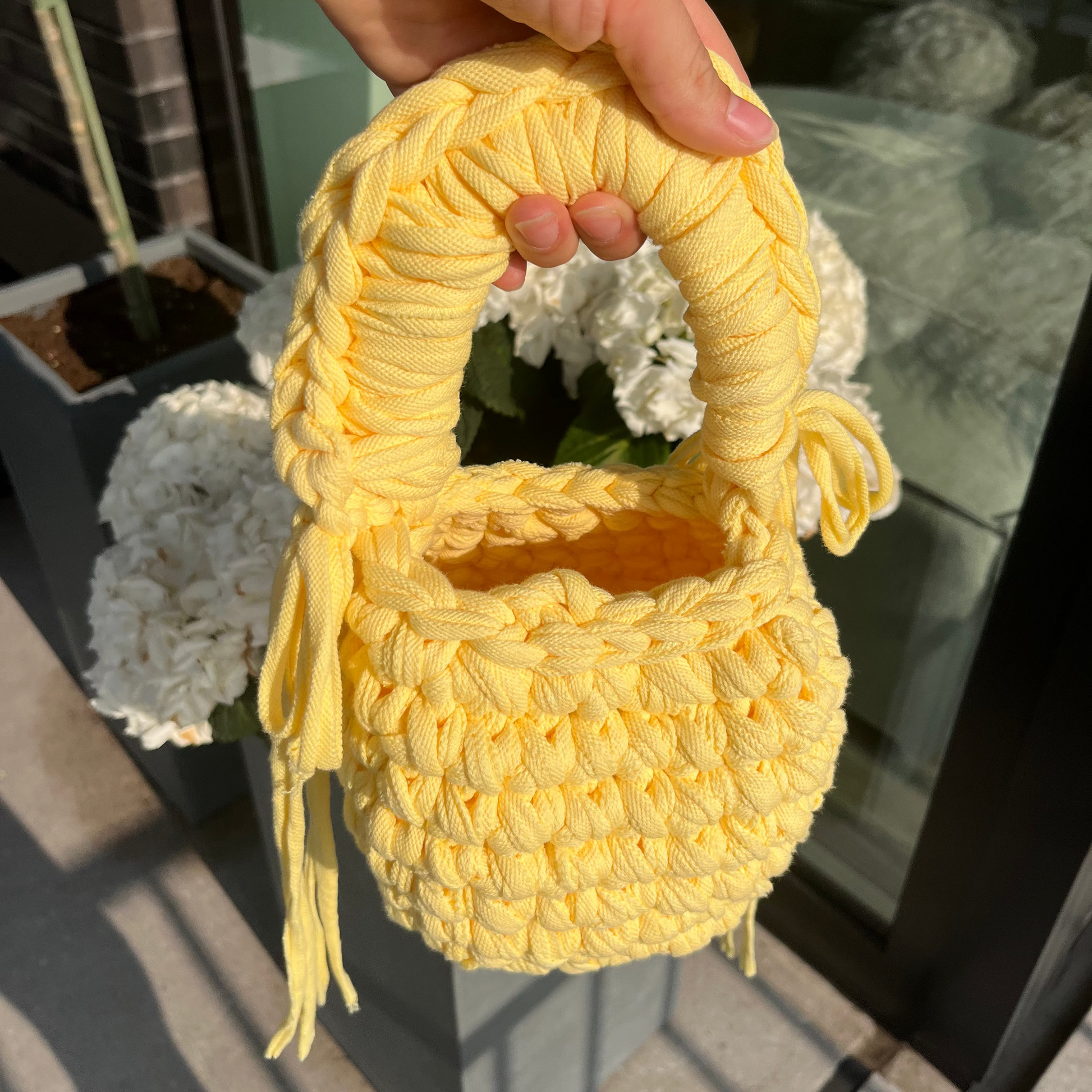 Yellow woven handbag held by a hand with a red bracelet, outdoor setting.